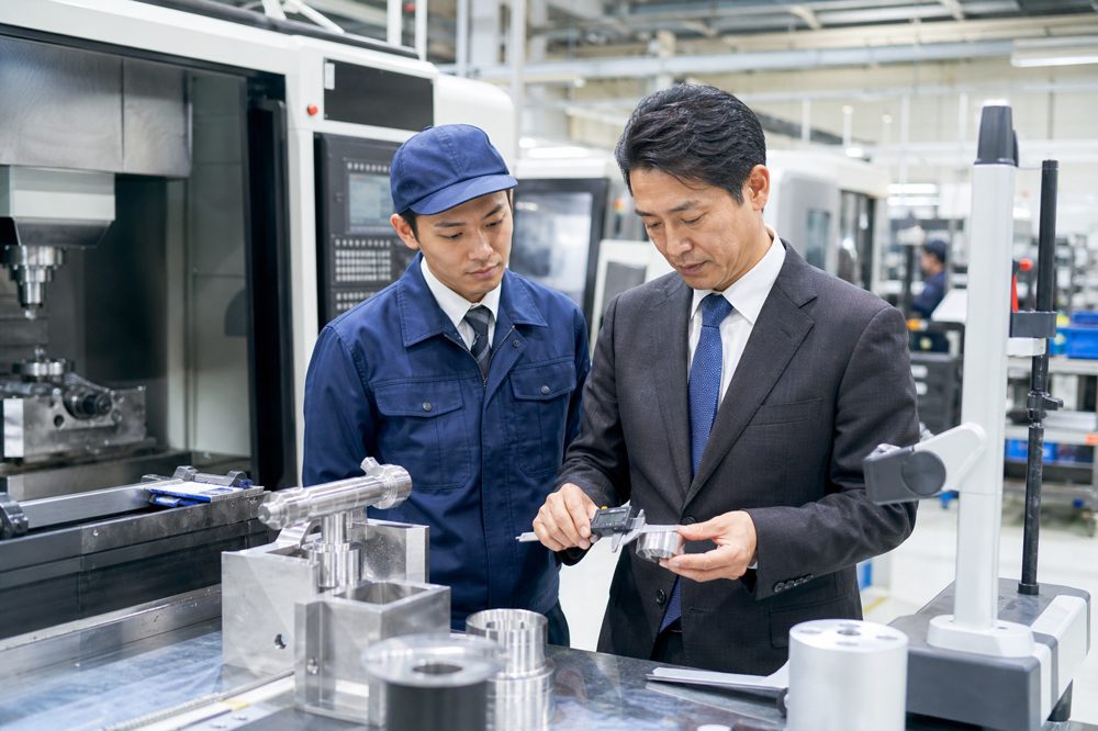 日本の清潔な金属工場でデジタル計測ツールを使いCNC加工機械を調べる現場監督とコンサルタント