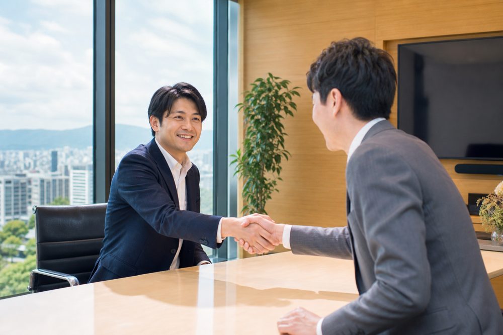 九州の明るい会議室で新しいビジネスパートナーと握手を交わし、満足した表情の中小企業経営者