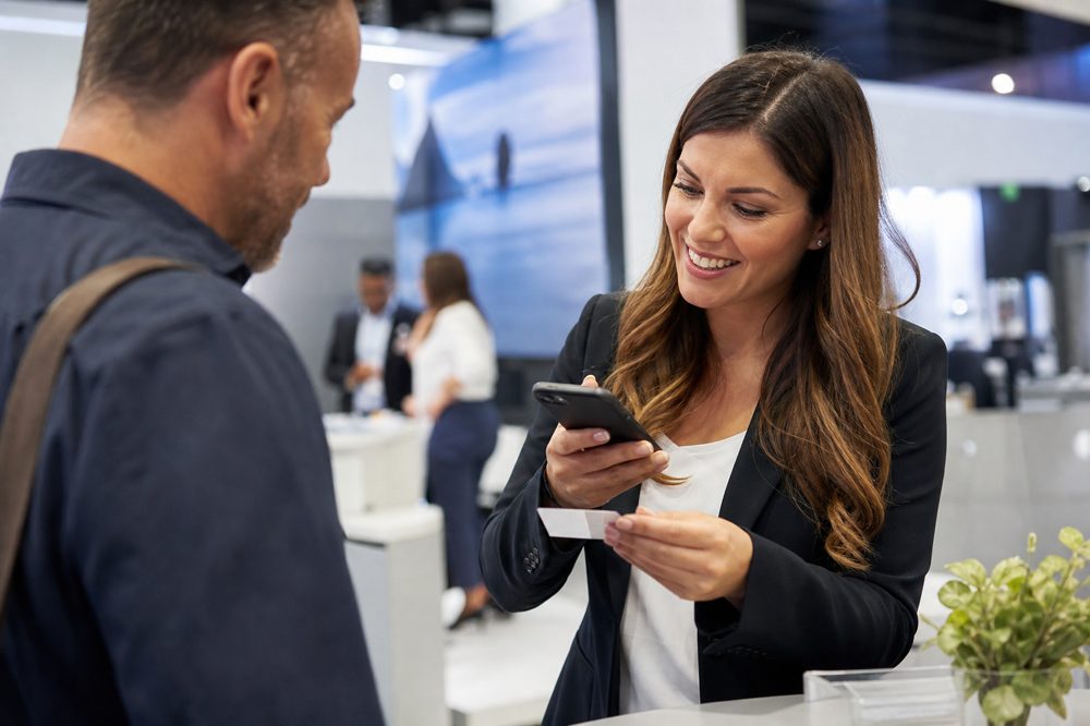 展示会ブースで来場者と話しながらスマートフォンアプリで名刺をスキャンする営業担当者