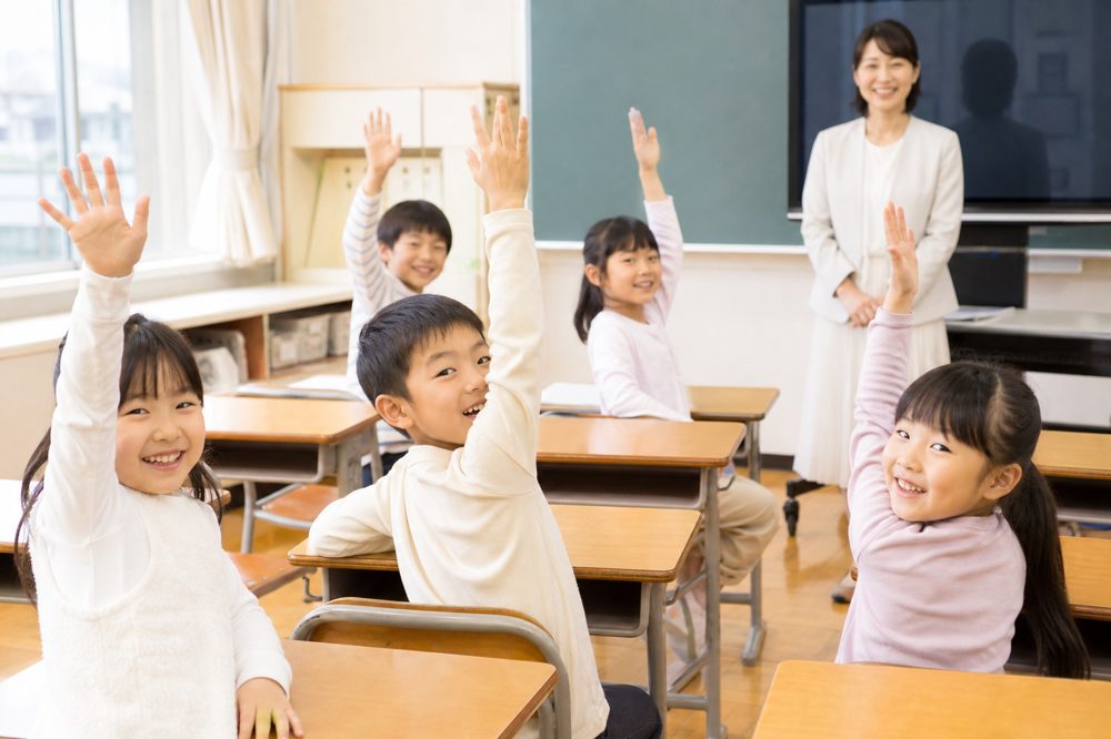 塾の授業で元気よく手を挙げる小学生たちと笑顔の先生