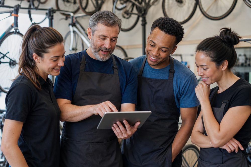 自転車店のオーナーとスタッフがタブレットを囲んで顧客管理アプリの使い方を学んでいる場面
